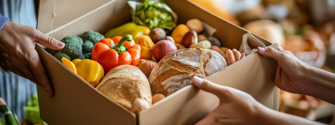 In a heartfelt act of giving, hands are shown passing a Thanksgiving meal box overflowing with fresh vegetables, bread, and canned goods